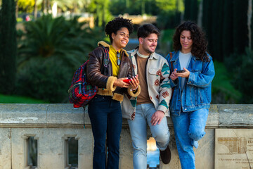 Fototapeta premium Three diverse friends standing on a bridge, smiling and looking at content on their smartphones, enjoying a city break together in a vibrant urban park