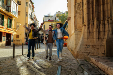 Three diverse friends holding hands, laughing, and jumping with arms raised while exploring an old european city street, sharing moments of happiness and adventure during their vacation © DusanJelicic