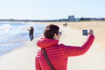 Woman in a red jacket taking a selfie near the shore on a sandy beach on a sunny autumn day
