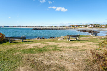 Empty clifftop benches facing a bay on a clear autumn day. A sandy beach lined with houses is visible in distance.
