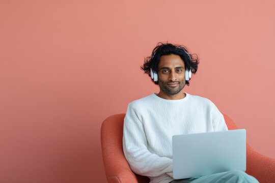 Male asian adult wearing headphones with laptop seated in an orange chair against a coral background for remote work and leisure