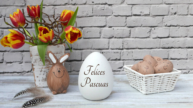 Un huevo de Pascua con la inscripci&oacute;n en espa&ntilde;ol "Felices Pascuas", un conejo marr&oacute;n y una cesta con huevos decorados frente a una pared de ladrillos blancos.