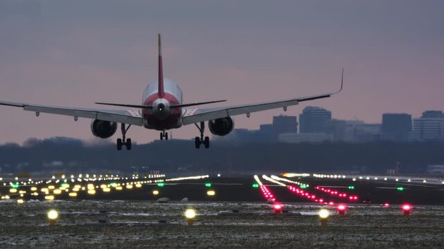 A passenger airplane descends, touches down, and rolls down an illuminated runway at twilight or dawn. Bright navigation lights guide the aircraft, with city buildings in the background