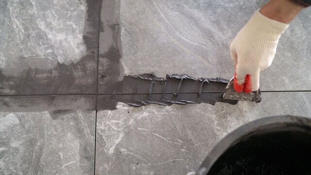 Close up of a worker's gloved hand using a trowel to apply dark grey grout between large stone-look floor tiles. Home renovation, construction, and DIY concept.
