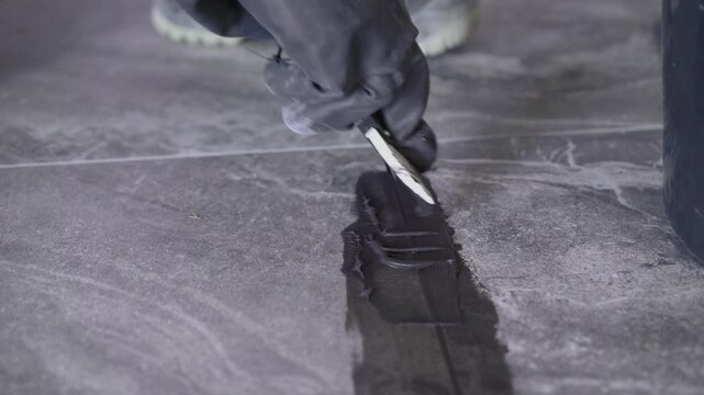 Close up of a worker's gloved hand using a trowel to apply dark grey grout between large stone-look floor tiles. Home renovation, construction, and DIY concept.
