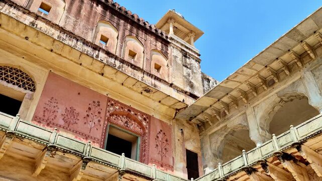 A bright daytime  showing a decorated inner wall of Amer Fort with faded pink floral paintings, arched windows, carved balcony railing, and upper watchtower structure under a clear blue sky.