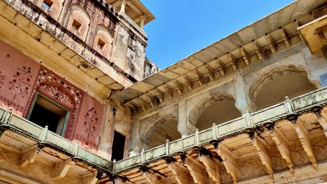 A bright daytime  showing an inner courtyard of Amer Fort with weathered sandstone walls, carved marble arches, painted floral doorway, and an upper balcony pavilion against a clear blue sky.