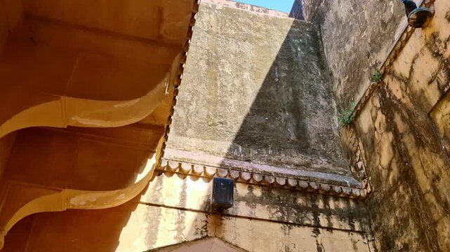 A bright daytime  showing narrow inner fort walls of Amer Fort with weathered yellow plaster, pigeons resting on stone holders, and a domed pavilion visible above against a clear blue sky.
