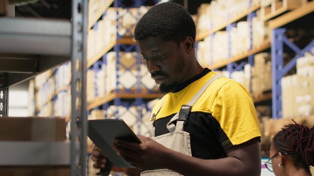 African american e-commerce worker scanning shipping labels numbers, registering awb and shipment details for the inventory records. Young man handling pallets distribution operations. Camera B.