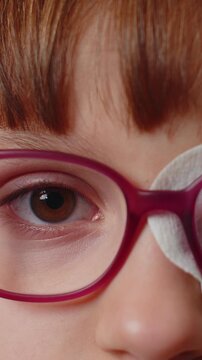 Close-up portrait of sick ill girl face looking at camera, having protective bandage plaster patch scratch on one eye. Injury, blind. Child kid in glasses. Medical eyesight vision therapy correction.