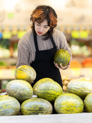 Fototapeta premium Young woman seller in apron puts fresh melon on display at vegetable market
