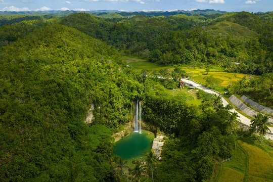 Dramatic aerial view of Camugao Falls in Balilihan, Bohol. Tall waterfall plunging into a deep pool within a massive limestone cliff amphitheater and lush forest.