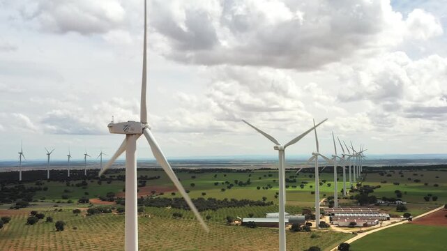 Drone shot of wind turbines generating renewable energy across vast farmland with green fields, farm buildings and cloudy sky, showcasing clean energy