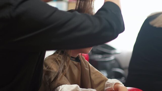 A cheerful little girl sits in a bright salon chair as she gets her first haircut. The stylist carefully trims her hair, creating a fun and memorable moment.