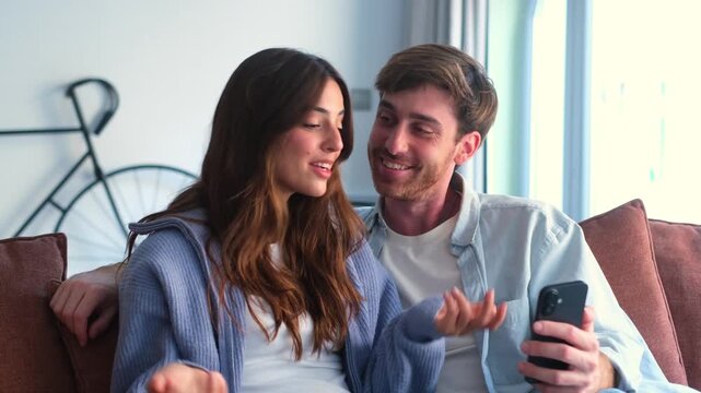 A young couple shares a joyful moment, looking at a smartphone on the couch. They react with surprise and laughter while watching content together, showcasing happiness and connection.