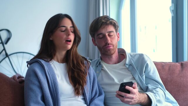 A young couple on a sofa looks at a smartphone, but the woman becomes visibly bored and frustrated with the device.