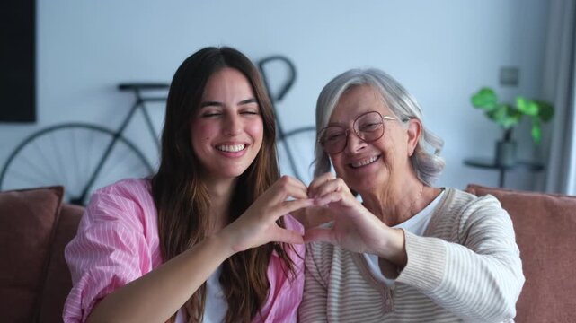 Smiling young and senior women form a heart with their hands, radiating love, family connection, and intergenerational happiness.
