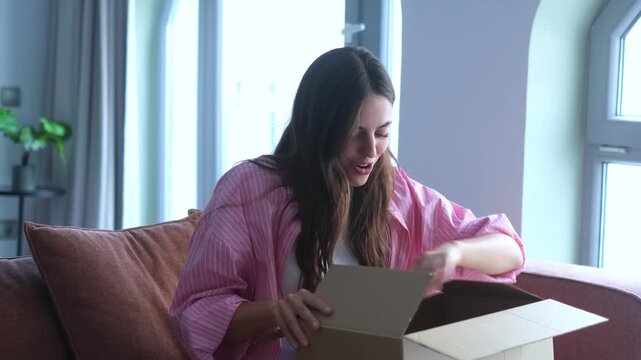 A young woman joyfully opens a cardboard delivery box at home, revealing a pleasant surprise inside. Her excitement and happiness are evident as she discovers the contents of her online purchase.