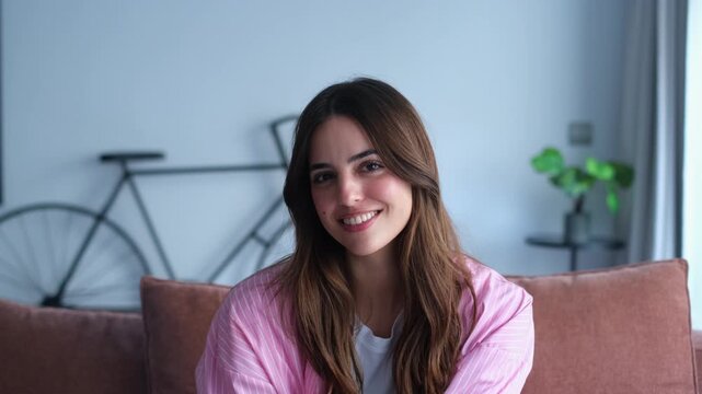 A beautiful young woman with long brown hair smiles genuinely at the camera, conveying happiness and positive emotions while relaxing comfortably at home.