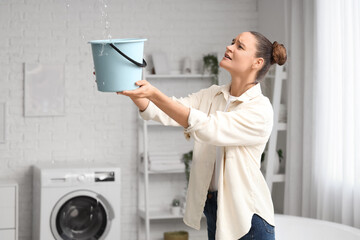 Worried young woman placing bucket under water dripping from ceiling in bathroom