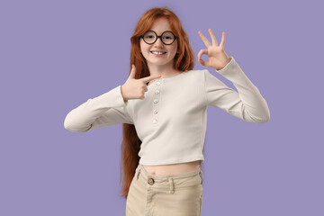Naklejka na ściany i meble Teenage redhead girl pointing at her OK gesture on lilac background