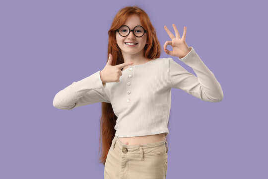 Teenage redhead girl pointing at her OK gesture on lilac background