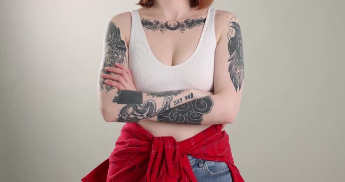 Beautiful woman with stylish tattoos, piercings and crossed arms on grey background. Camera moving down