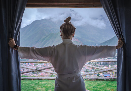 Woman tourist wearing a white bathrobe looking through a window at the Sacred Valley of the Incas in Peru. Travel and relaxation concept in a luxury accommodation, representing tourism, wellness.
