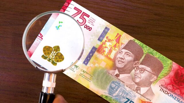 A magnifying glass inspects a commemorative 75,000 Indonesian Rupiah banknote. The video highlights portraits of founding fathers, security watermarks, and festive colors celebrating independence.