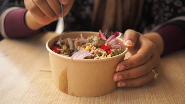 Close up view of woman enjoying healthy tuna salad with fresh vegetables in disposable paper bowl eating with fork at wooden table