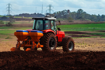Fototapeta premium Red tractor with a seeder plowing a field in a rural landscape, with power transmission towers in the background, symbolizing modern agriculture and energy infrastructure. 