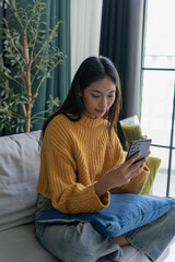 Obraz premium Asian woman in sweater using smartphone and drinking green vegeteble juice