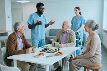 Obraz premium Wide shot of senior men and senior woman socializing with caregivers in care home, playing chess and drinking coffee. Supporting elder care, retirement living, healthcare marketing