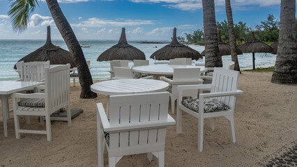 Seaside outdoor cafe. White plastic tables and chairs with cushions on the sand. Palm trees, straw...