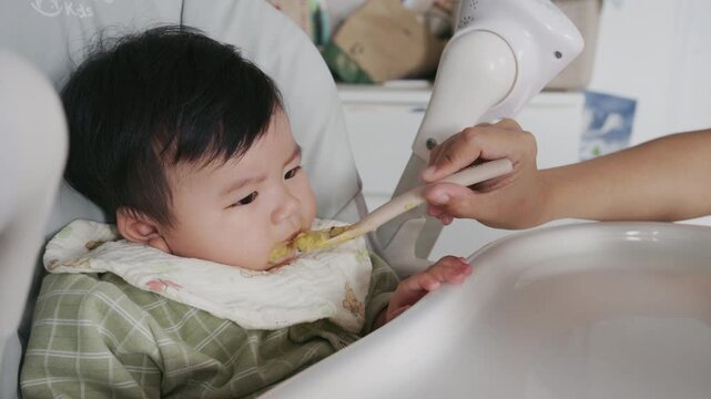 4K Asian mother feeding her six month old baby boy with a spoon at home in morning.
