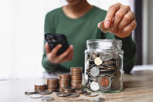 Close-up of a woman saving money by putting coins into a glass jar while checking her smartphone. Representing personal finance, budgeting and financial planning.