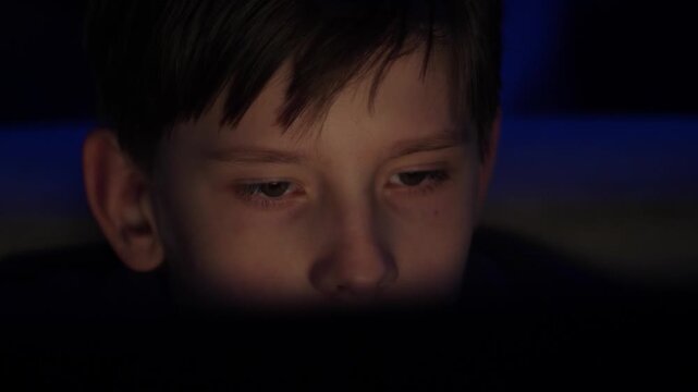 A boy is looking at a computer monitor in the dark. Close-up portrait with blue background lighting
