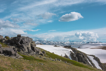 Scenic view to sunlit grassy hillside with rocky outcrops against snowfields on rocks and big snowbound rock mountain range far away under clouds in blue sky. High snow mountains in changeable weather © Daniil