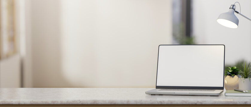 Blank screen laptop with book and lamp aside potted plant on marble counter table in white wall room