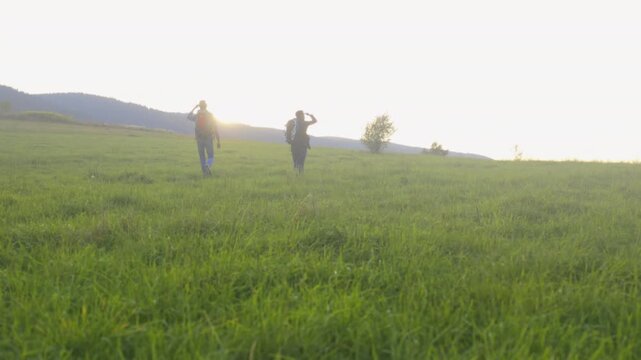 Sunlit meadow adventure with backpacks, Two adventurers with backpacks walk through radiant grass during sunset hour