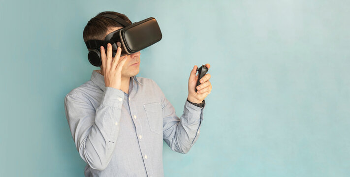 Young smiling man in VR glasses playing video game with joystick on a blue background, siting on the couch while having virtual reality experience, copy space