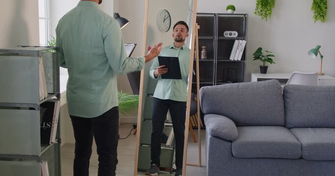 Man holds a clipboard, preparing for rehearsal and presentation, checking reflection in a mirror. Man rehearses presentation in front of mirror. Employee prepare for job conference at home