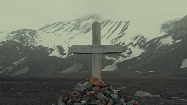 Weathered wooden cross with engraved inscription stands over stone-piled grave at Whalers Bay cemetery, Deception Island, Antarctica, with misty snow-streaked volcanic mountains behind