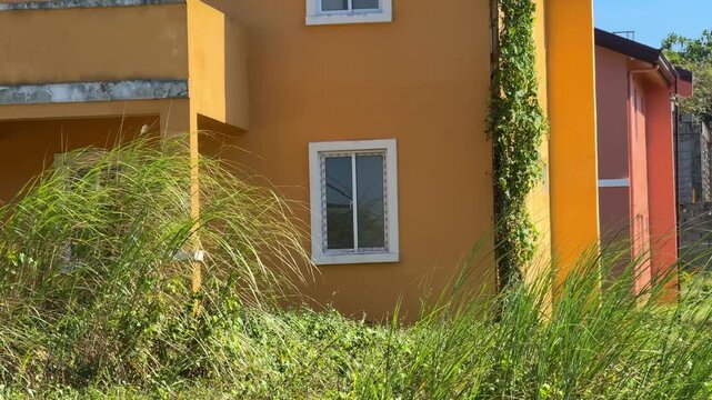 Ground level corner view of finished unoccupied house, tall grass up to walls at Camella Homes Alfonso Cavite Philippines