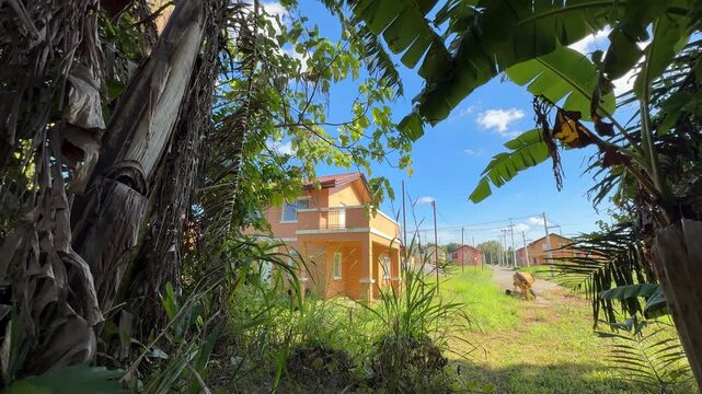 Ground level front drive view of finished unoccupied house, tall grass encroaching walls, old construction equipment nearby.