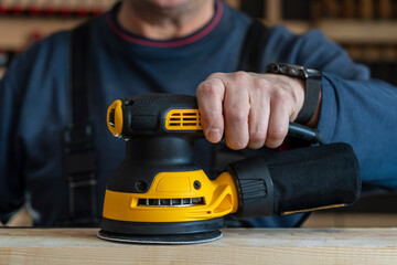 Craftsman sanding a wooden beam with an electric orbital sander in workshop. Professional woodworking and DIY home renovation concept. Carpenter using professional random orbit sander on wood © OlegD