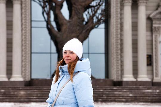 A young woman, warmly dressed for winter, pauses in Kazan. Snow dusts the steps of a grand building, framed by imposing columns, as she gazes into the distance.