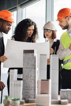 A diverse group of architects and engineers collaborate, examining building plans and a miniature architectural model in an office setting