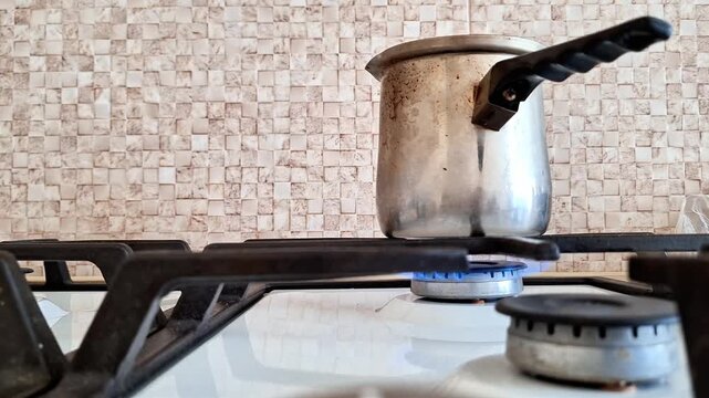 Metal pot with black handle heating on a lit gas stove burner, set against a beige tiled wall, illustrating everyday cooking, kitchen utility, and household food preparation