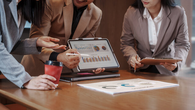 Smiling business team collaborating over documents in modern office with data charts on screen, discussing strategy and analytics during meeting.